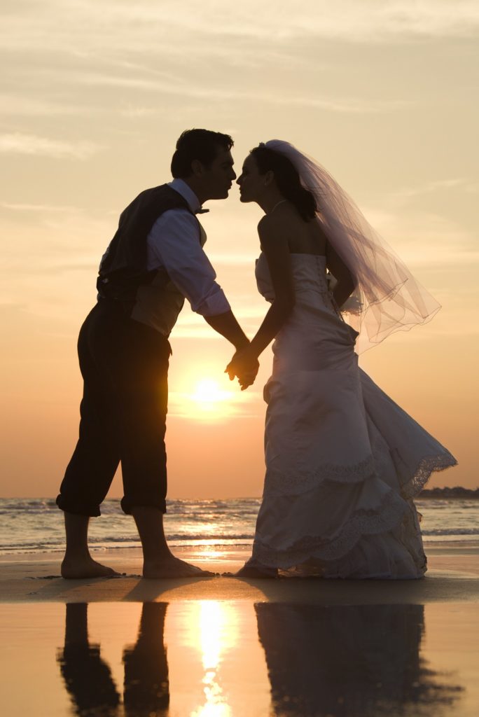 Caucasian mid-adult bride and groom holding hands and kissing barefoot on beach at sunset.