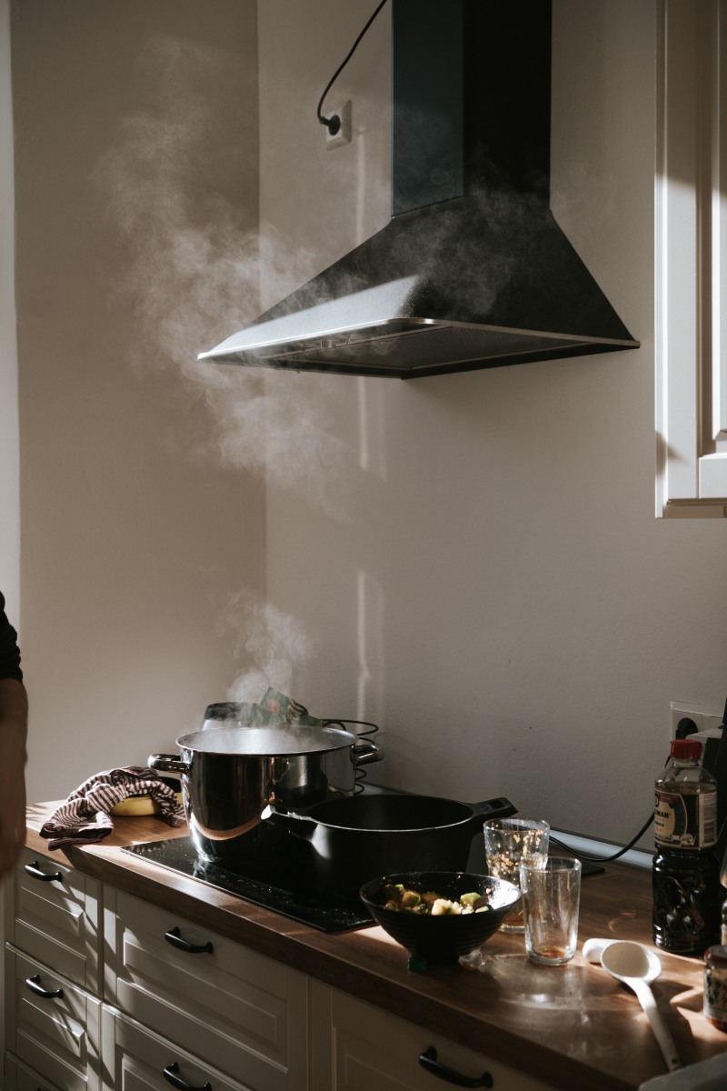 Kuchnia indukcyjna, na której stoi garnek z gotującą się wodą z którego paruje woda. Nad garnkiem znajduje się okap kuchenny.