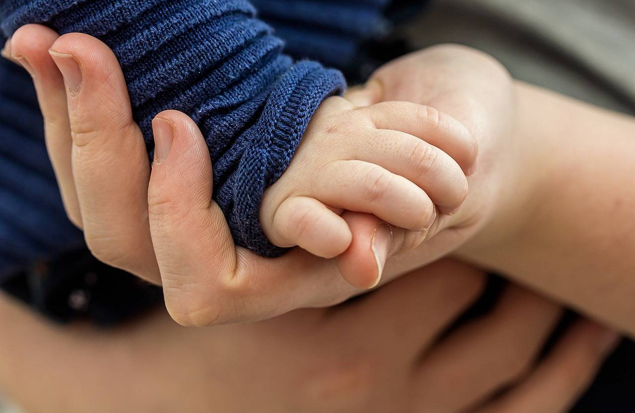 dłoń niemowlaka w granatowym sweterku opierająca się na dłoni osoby dorosłej