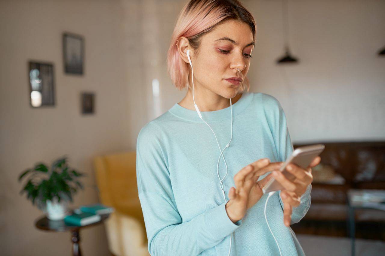 Serious young female smm manager with facial piercing working distantly using cell phone, promoting brand and expanding target audience via social networks, listening to music in earphones
