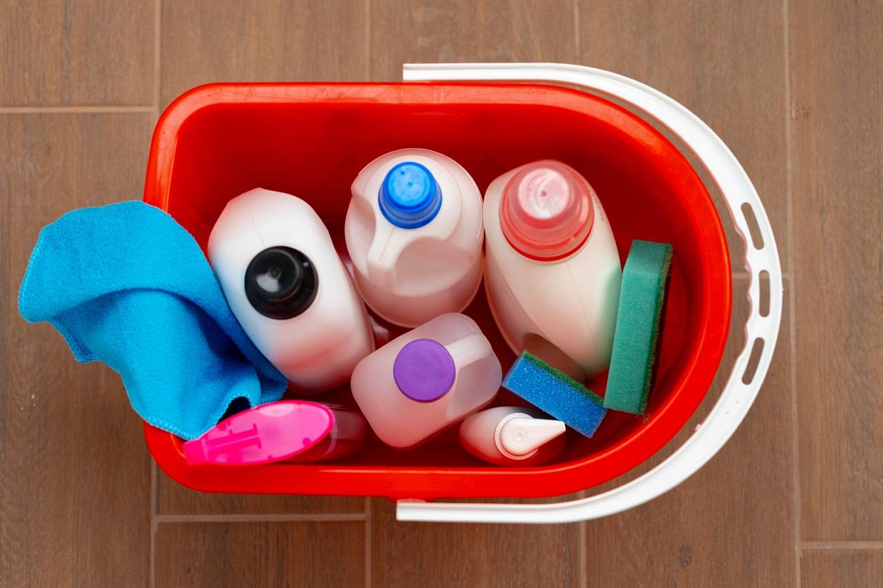 Various household cleaning detergents and bottles in a plastic bucket on the floor