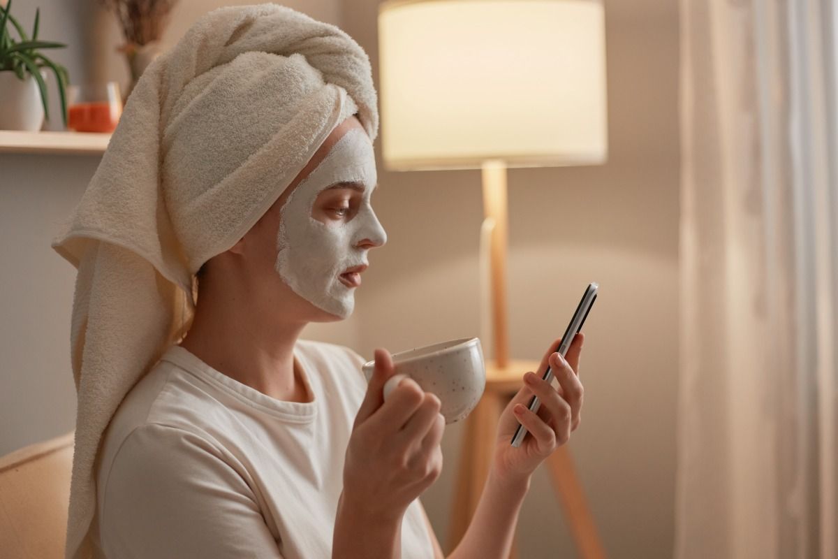 Side view photo of beautiful young adult female with clay mask on her face, woman in towel on head drinking coffee, holding cup and phone in hands, sitting on cough in her living room