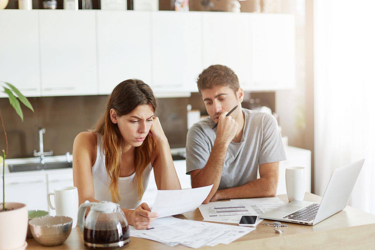 Puzzled female and male sitting together at table in cozy kitchen, studying papers or documents of their family business and working with modern laptop. People, finances, domestic budget concept