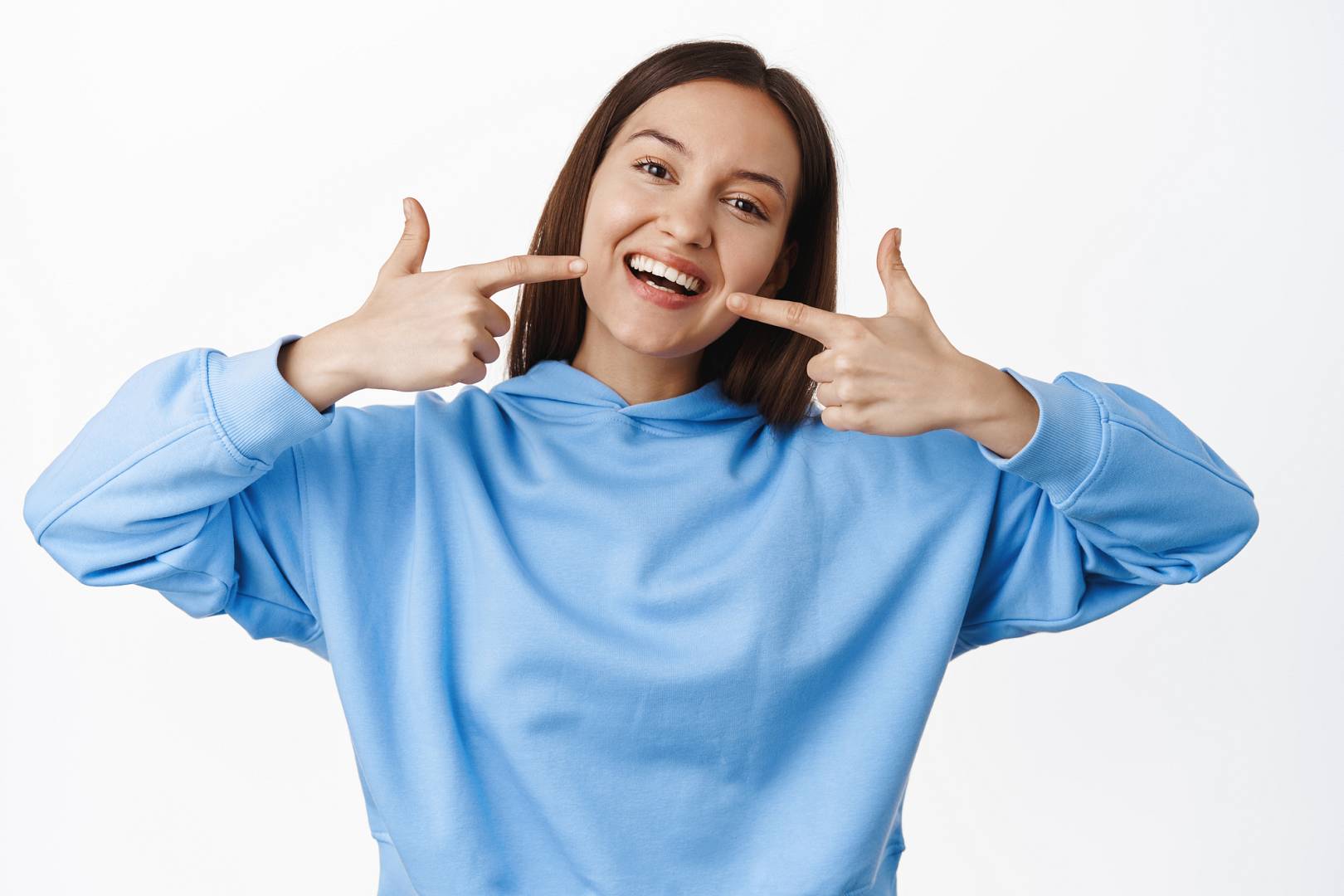 Happy girl pointing fingers at white perfect smile, straight whitened teeth, standing in hoodie against white background